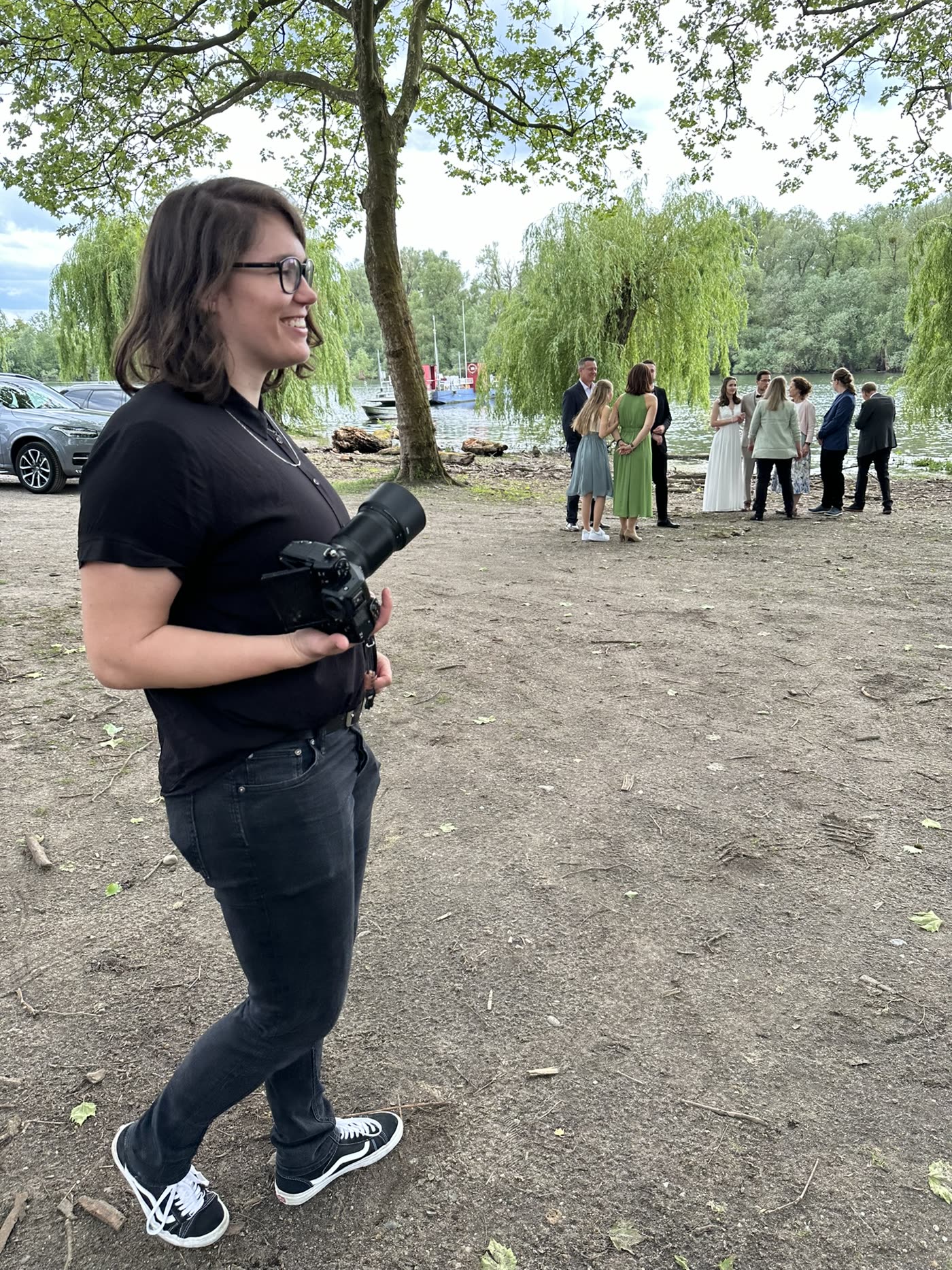Janet steht mit ihrer Kamera am Ufer. Dahinter wartet eine Hochzeitsgesellschaft.