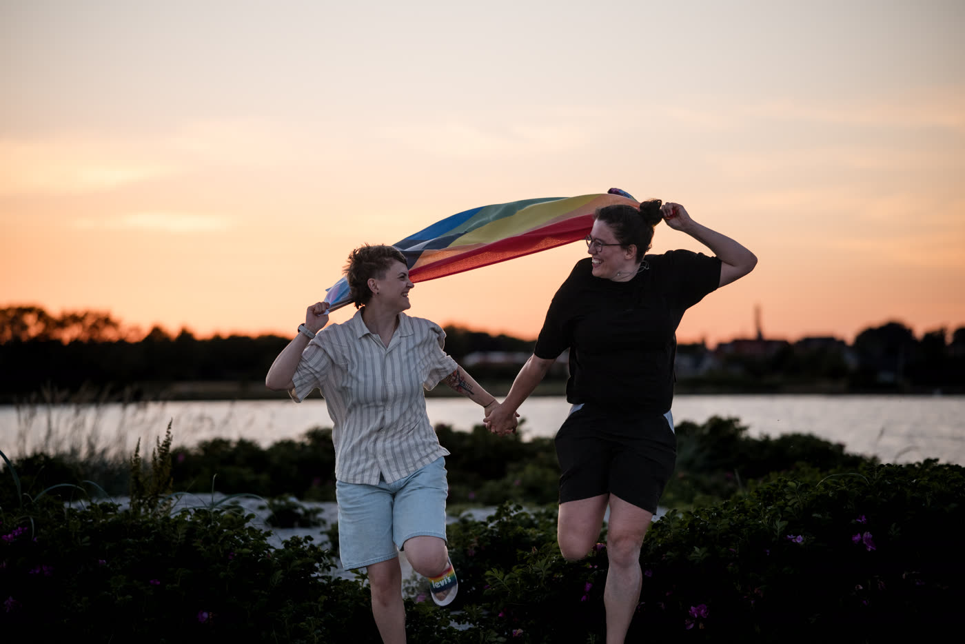 Zwei Menschen gehen bei Sonnenuntergang Hand in Hand unter einer Regenbogenfahne.