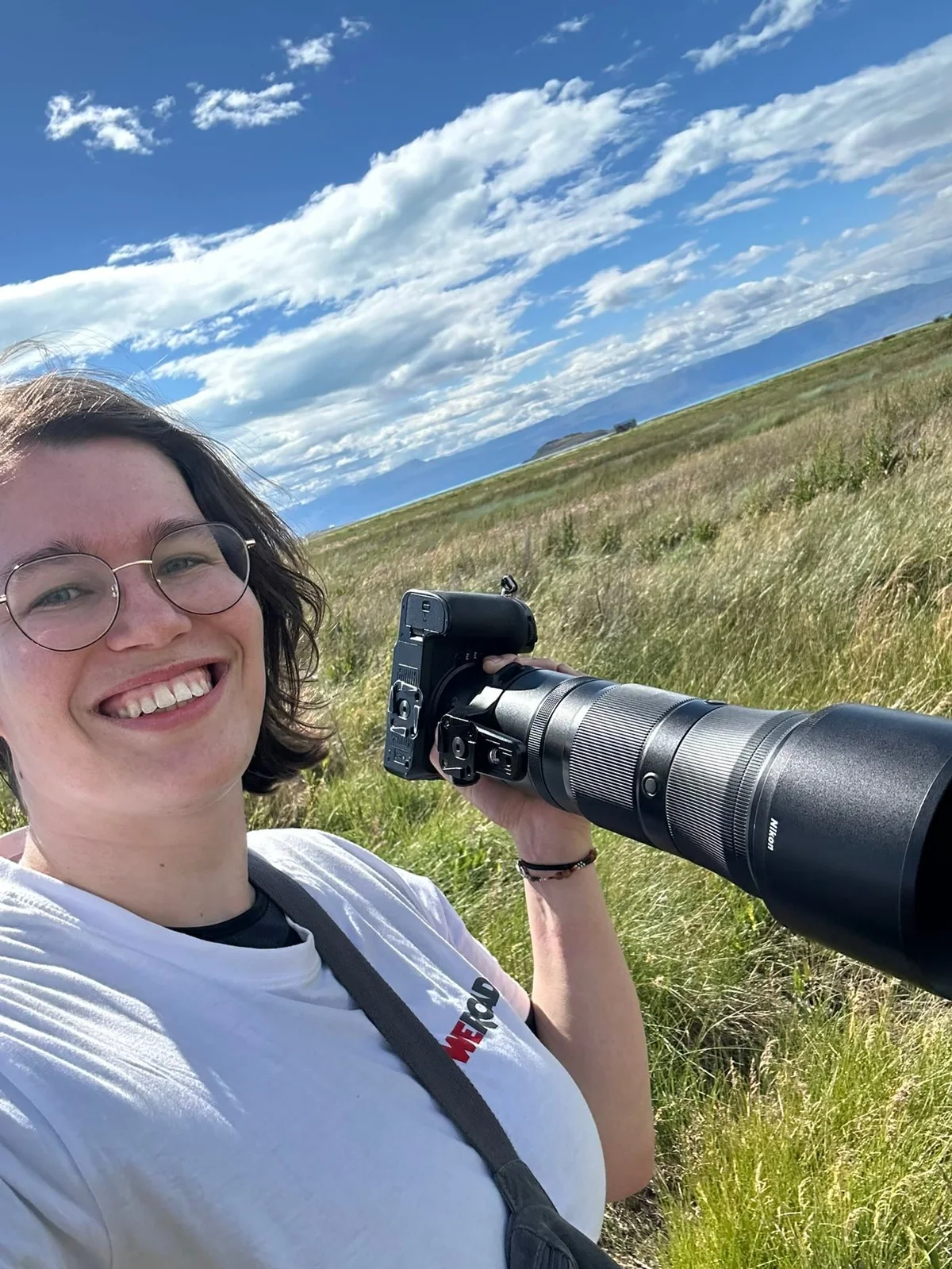 Janet laughs with her camera and a large telephoto lens in a meadow.