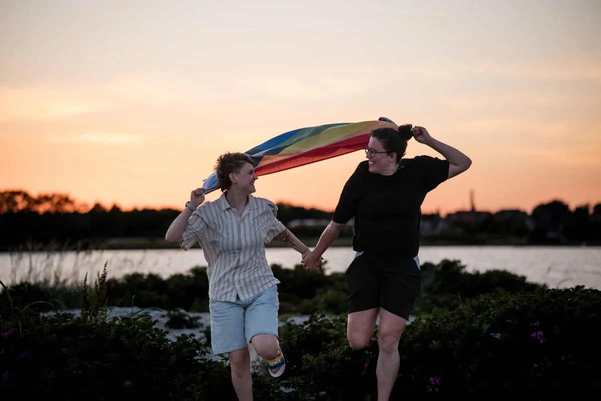 Two people walk hand in hand beneath a rainbow flag at sunset.