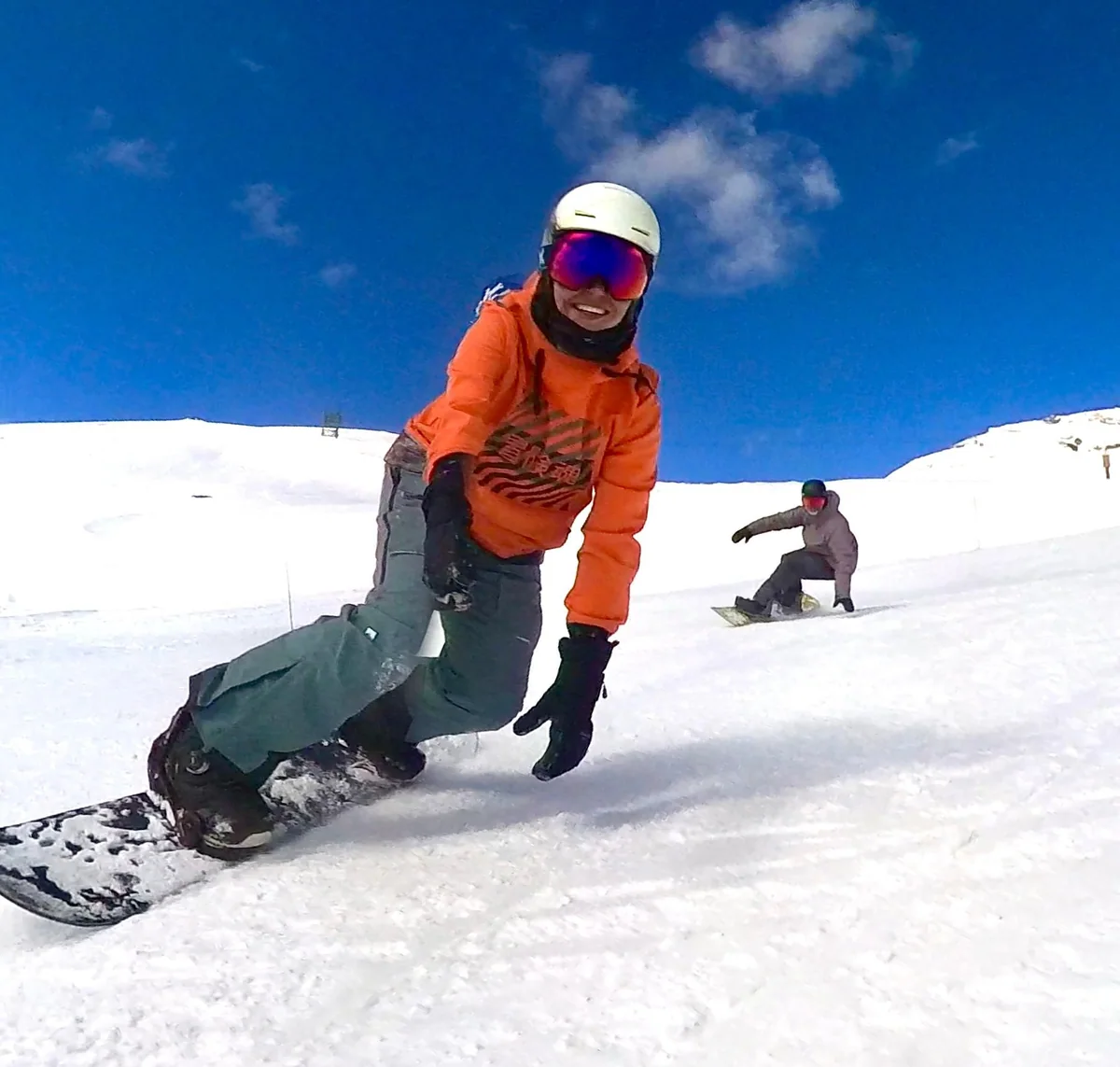 Janet laughs while snowboarding downhill in an orange jacket.