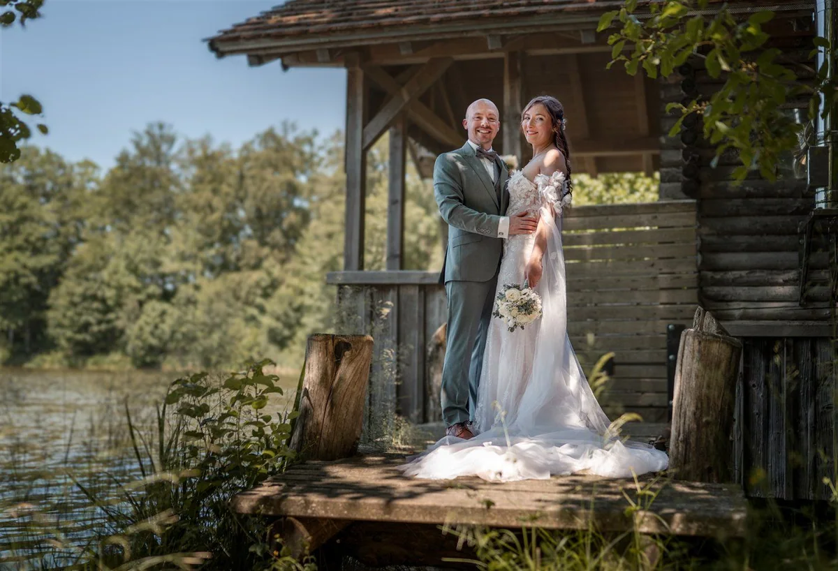 A couple stand on the jetty of a wooden boathouse by the lake.