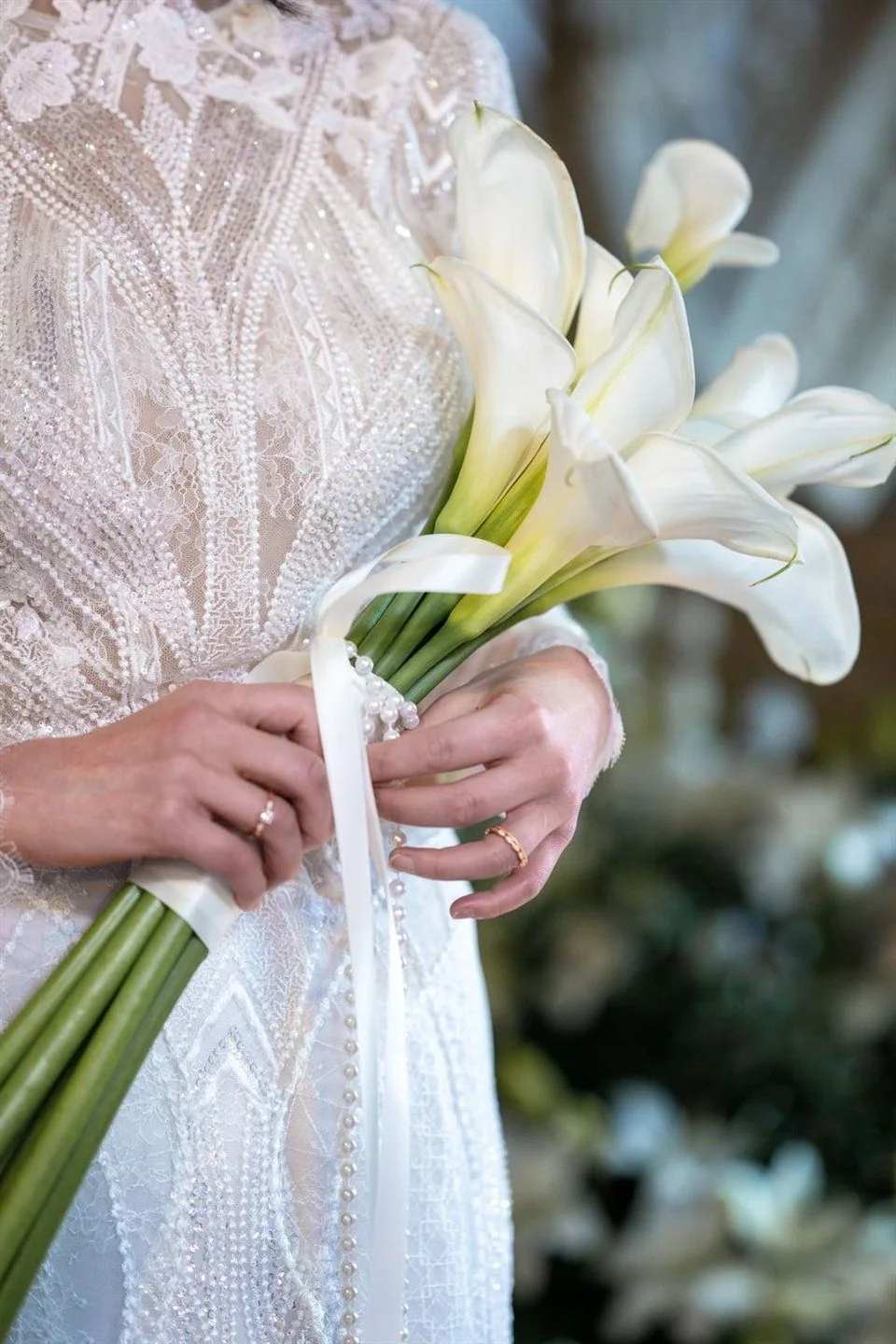 A bride holds white calla lilies in front of her lace dress.