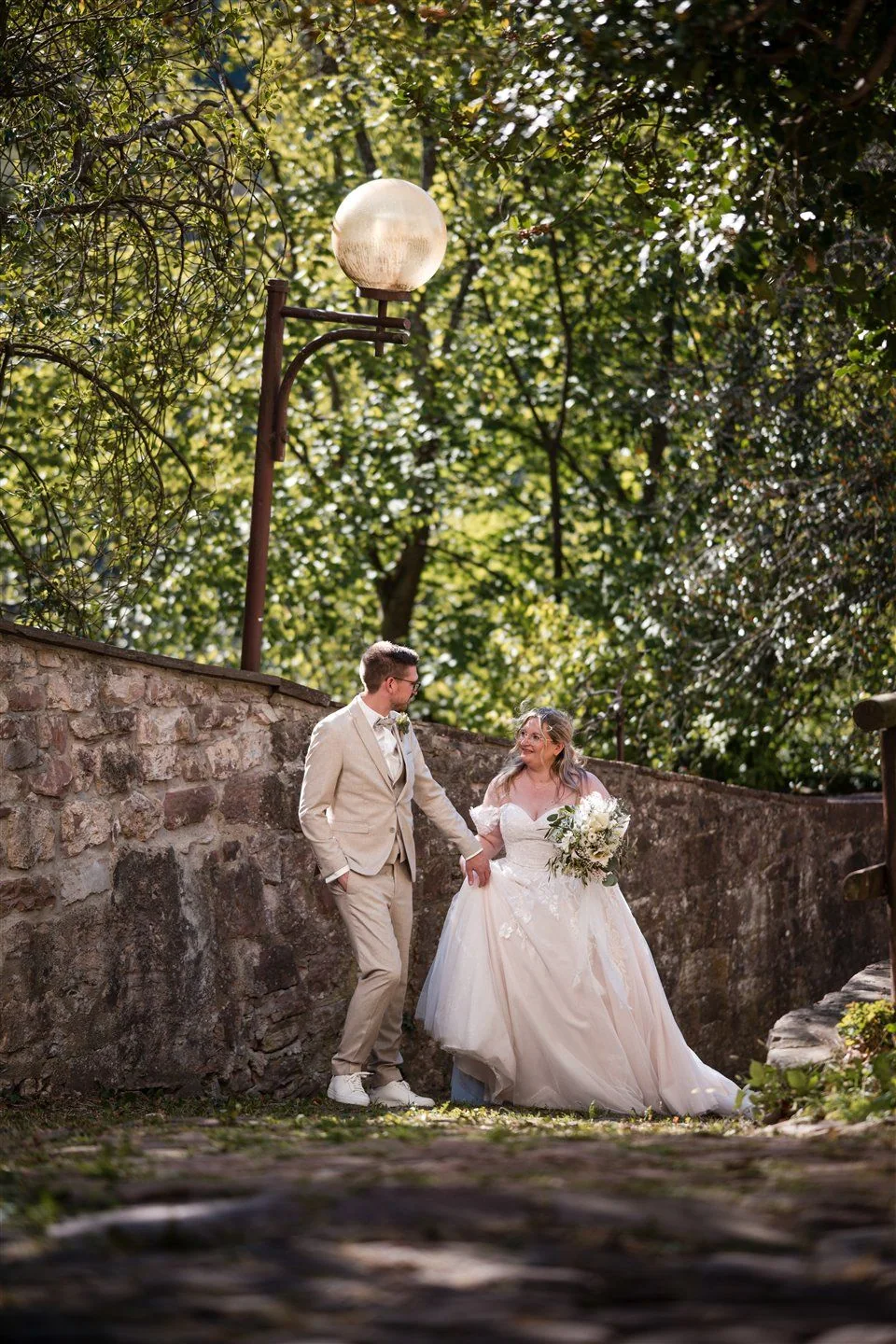A wedding couple walk beneath a lantern beside an old stone wall.