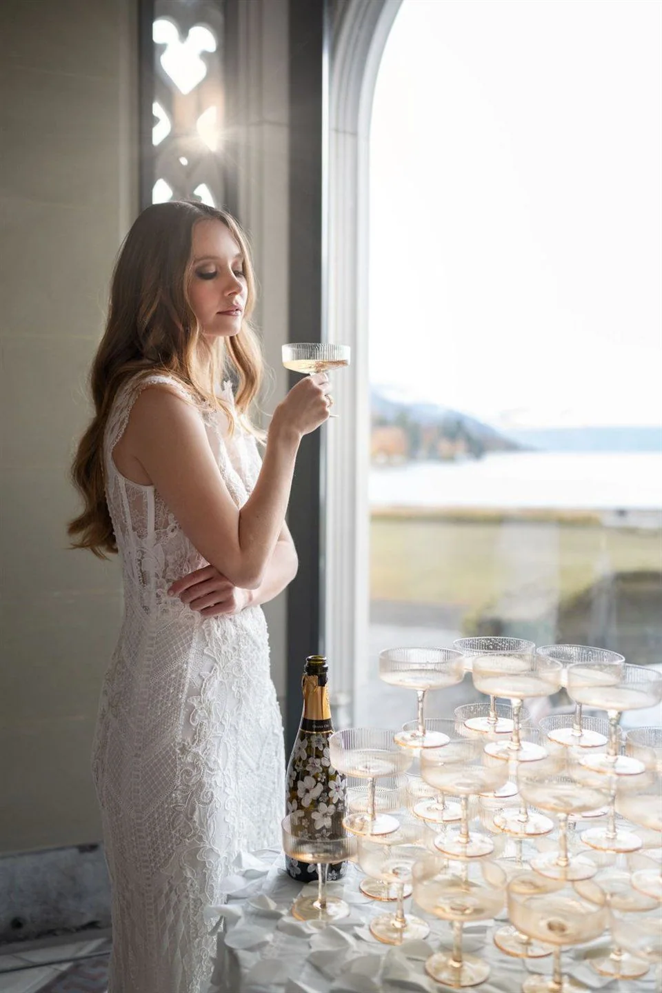 A person in a wedding dress stands by a window with a glass.