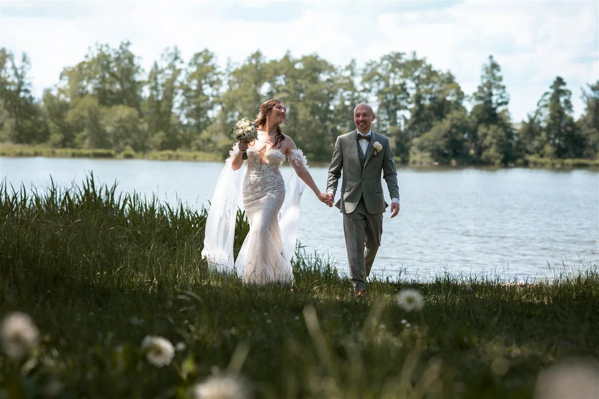 A couple walk hand in hand through tall grass beside a lake.