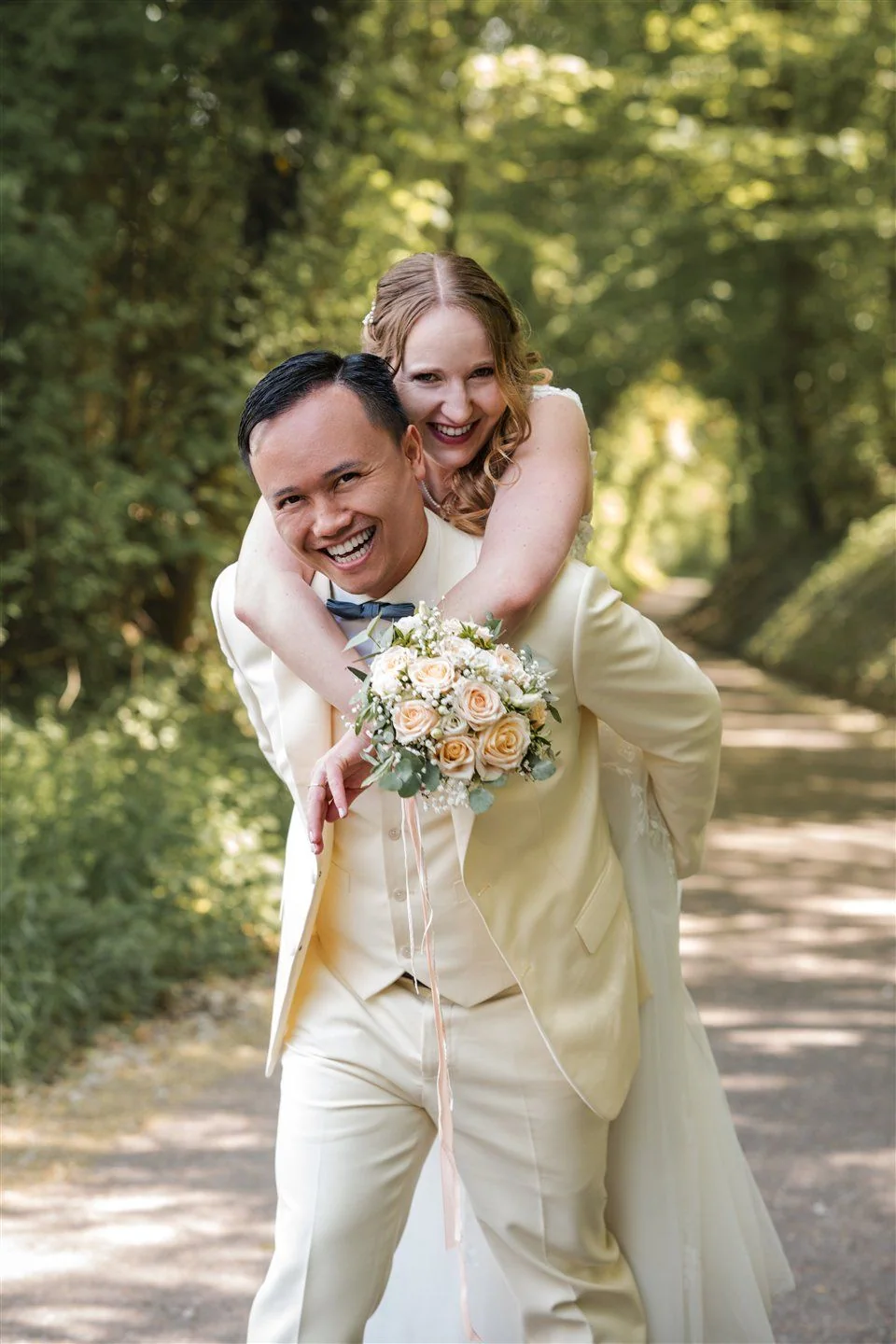 A bride laughs as she is carried piggyback along a forest path.