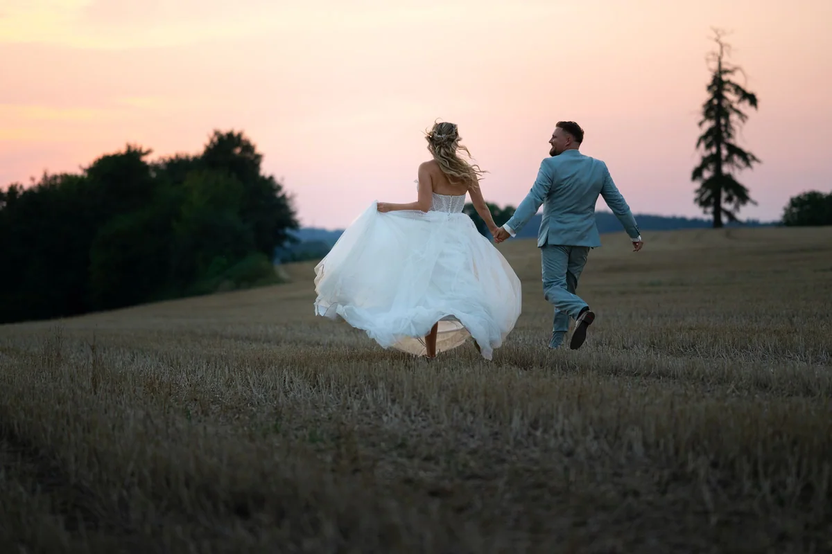 A wedding couple run hand in hand across a field at sunset.