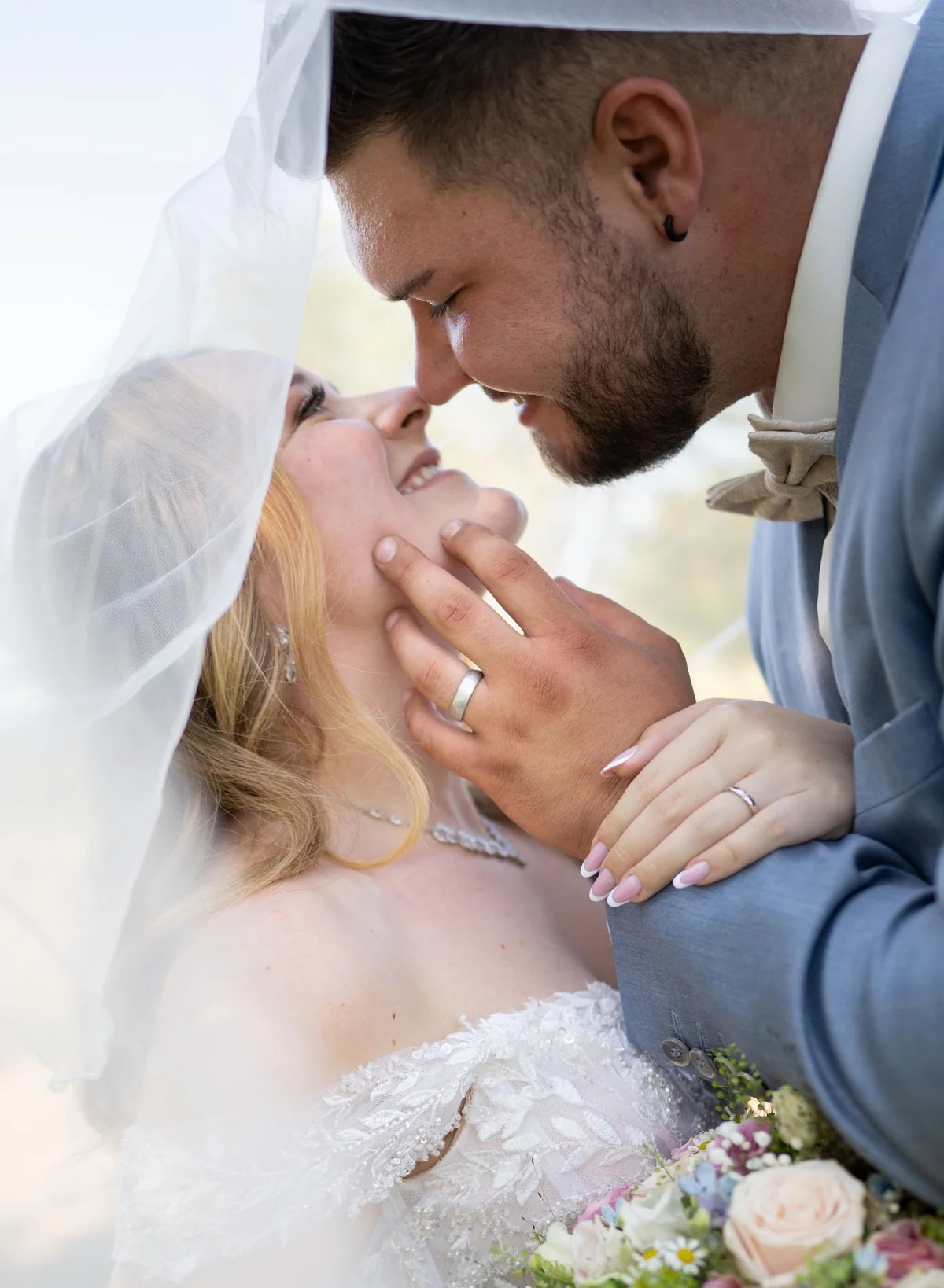 A wedding couple touch foreheads beneath a light veil.