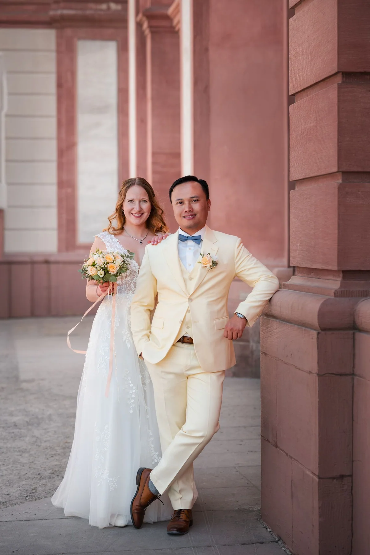 A wedding couple lean smiling against a pink wall.