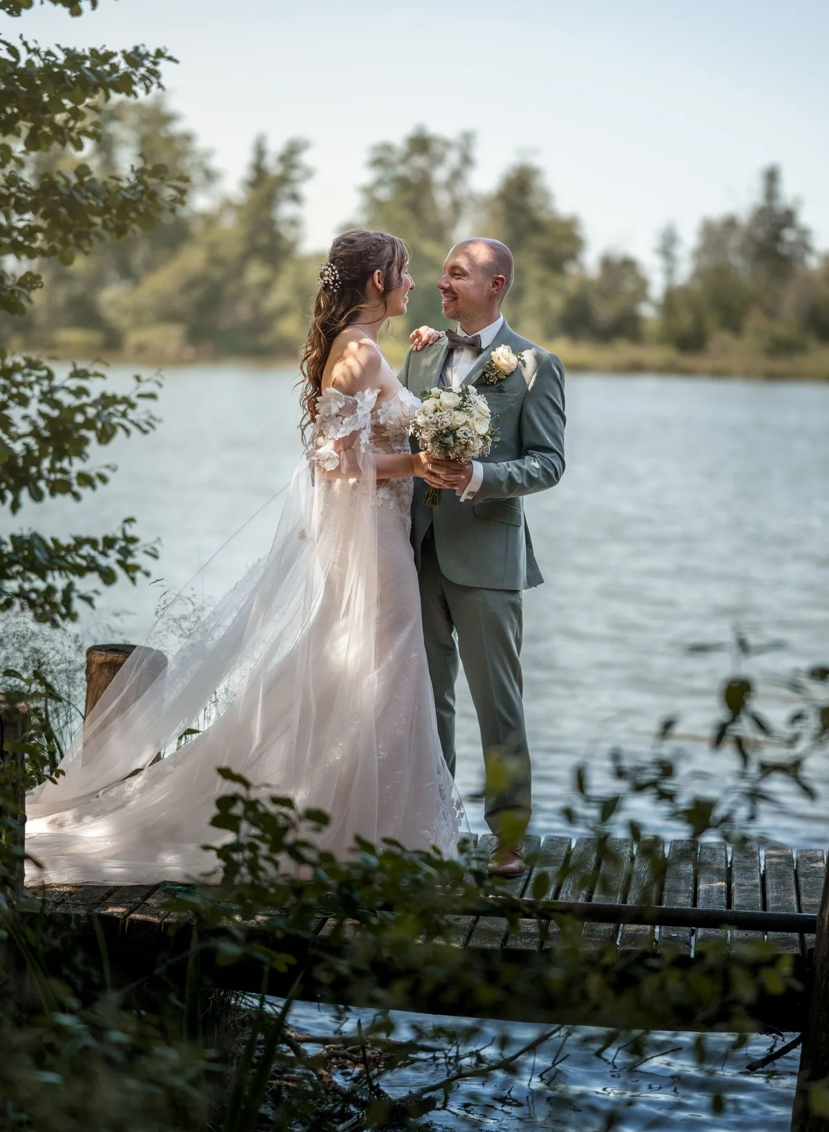 A wedding couple stand close together by the lake.