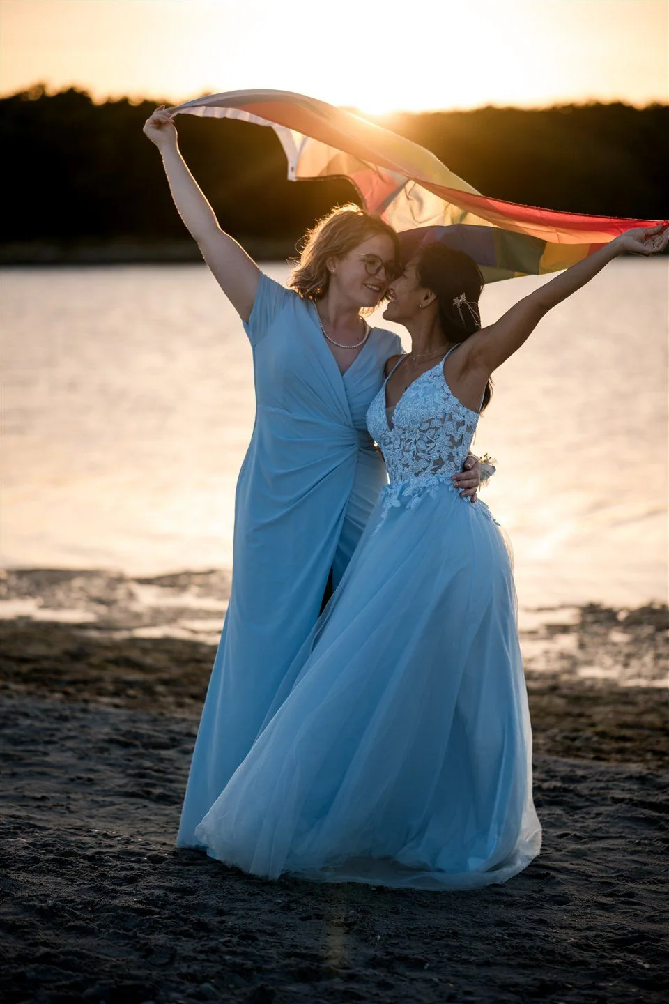 Two brides hold a rainbow flag above themselves towards the evening sun.