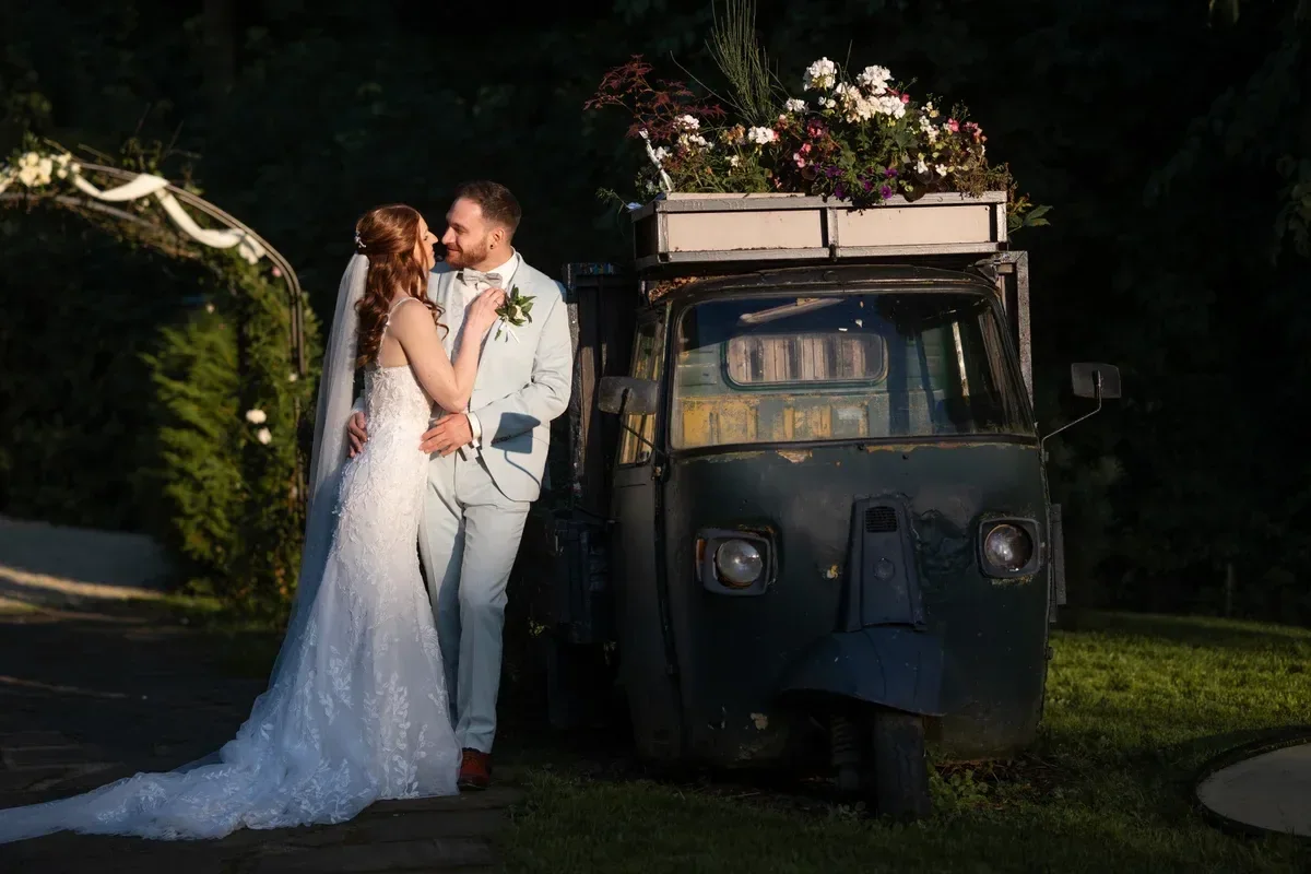Das Brautpaar steht abends neben einem kleinen Getränkewagen mit Blumen, die Sonne fällt seitlich auf die beiden.