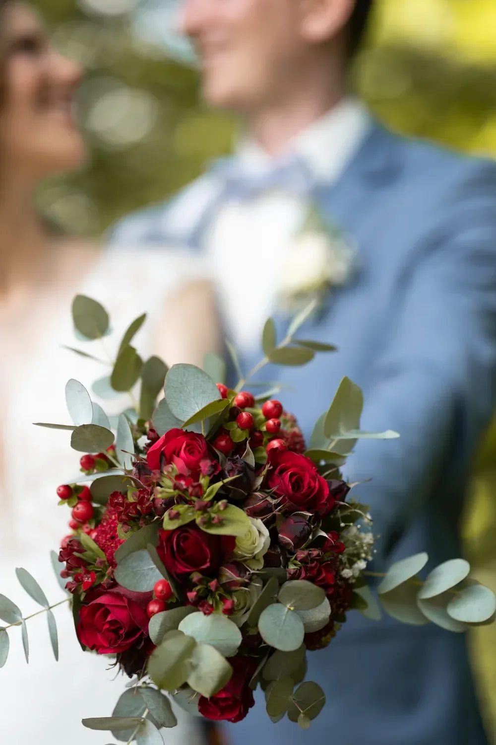 Ein Strauß aus roten Rosen und Eukalyptus, dahinter unscharf das Brautpaar.
