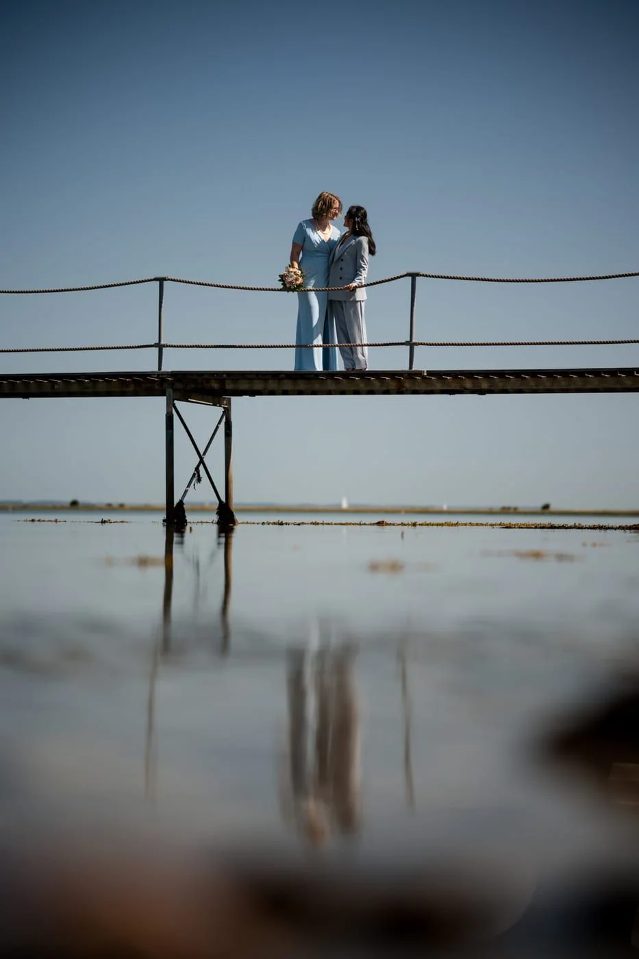 Two brides stand close together on a wooden jetty above the water.