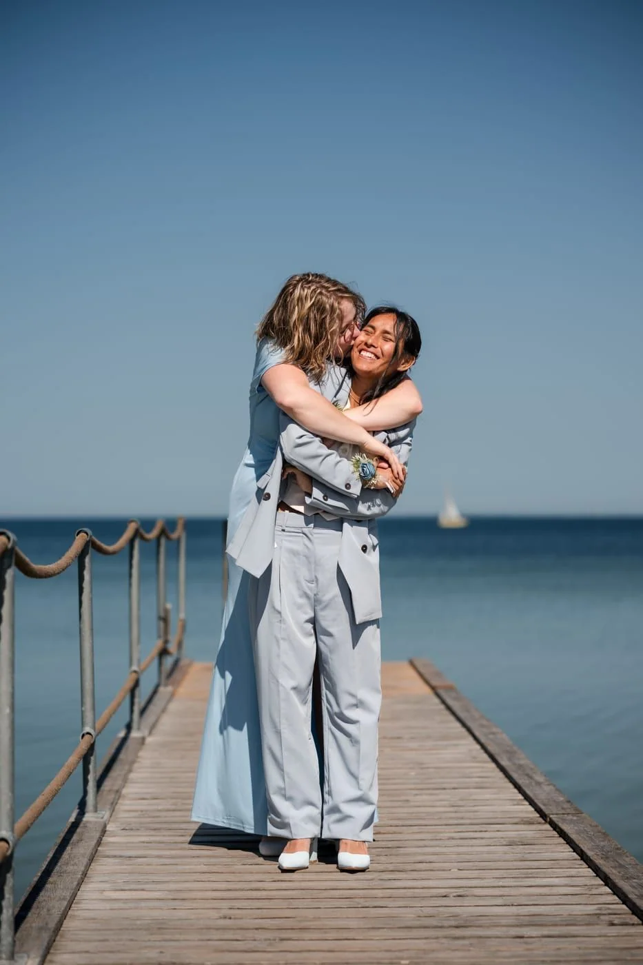 Two women in light coloured suits embrace laughing on a jetty by the sea.