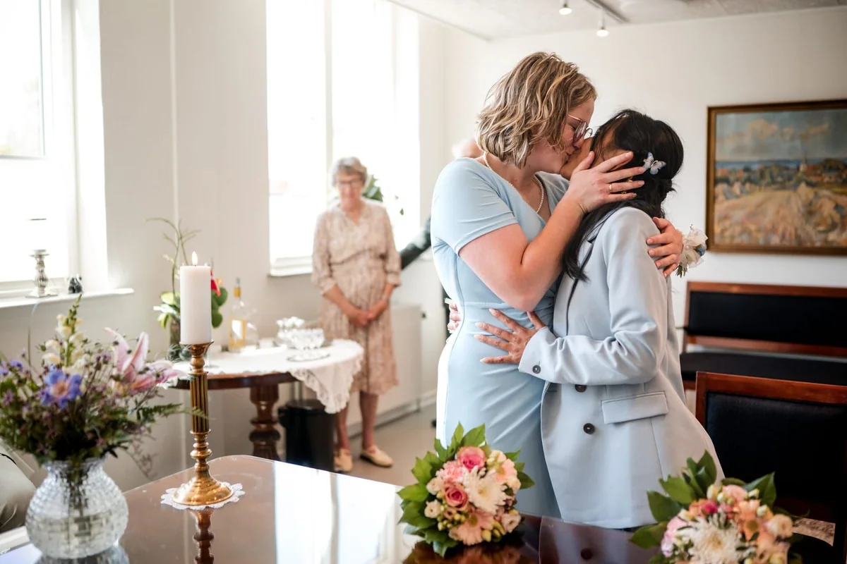 The couple kiss during their civil ceremony as a guest watches in the background.