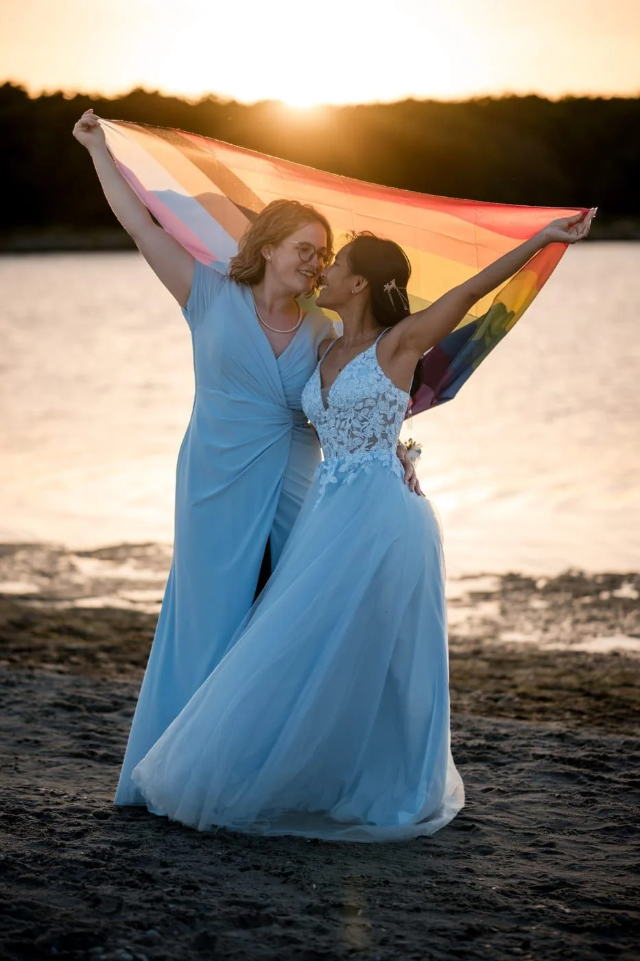 The couple hold a Progress Pride flag above them on the beach at sunset.