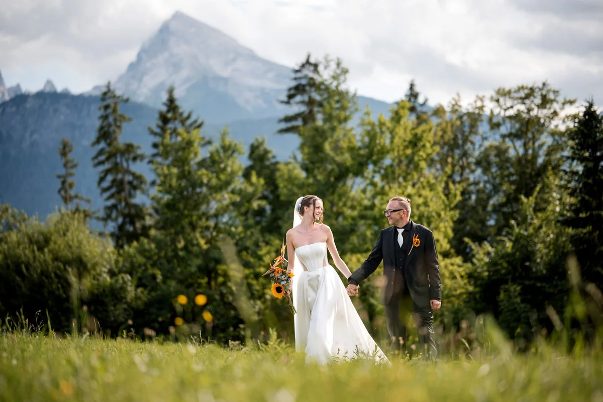 The wedding couple walk hand in hand through a meadow.