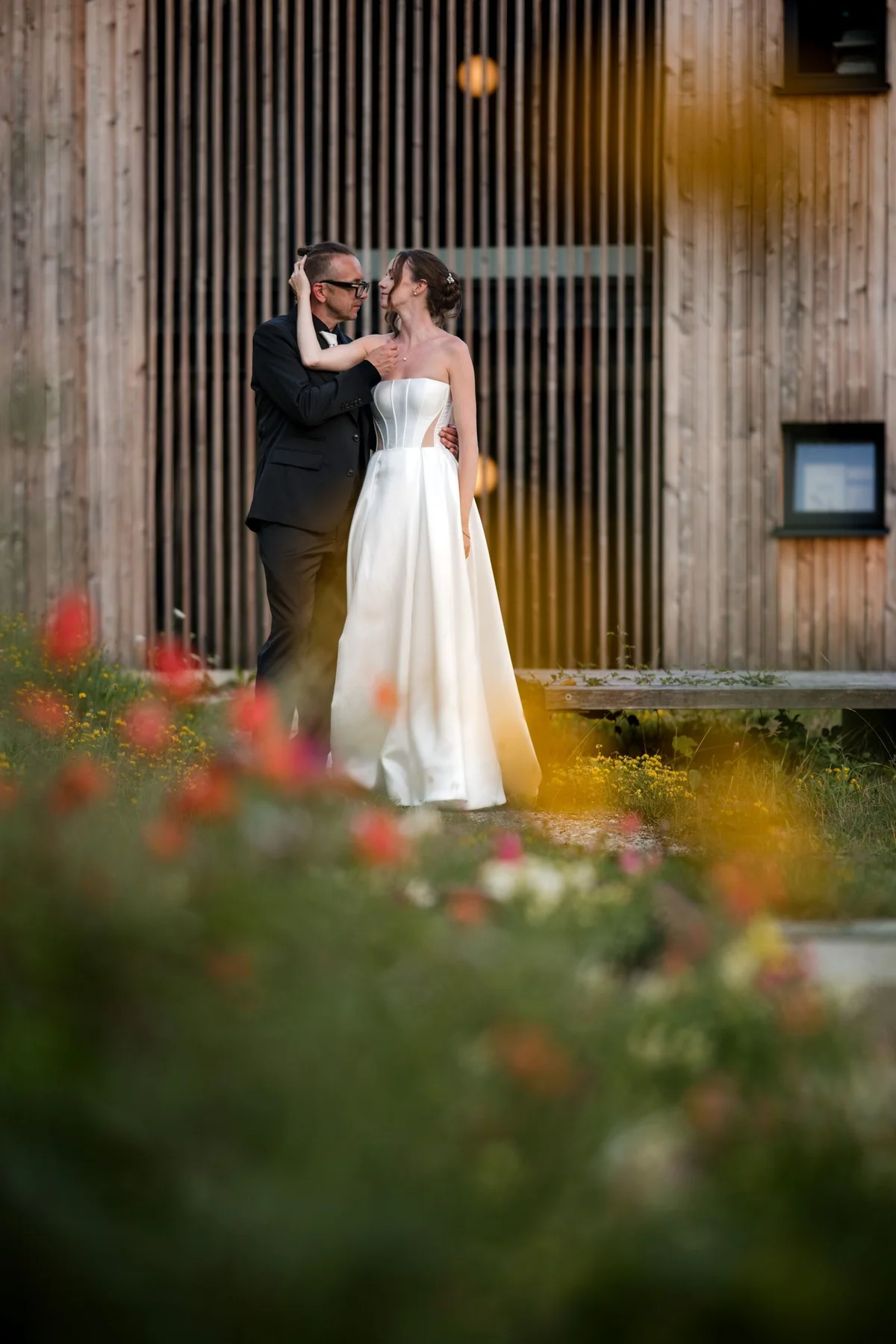 The couple embrace in front of a wooden facade.