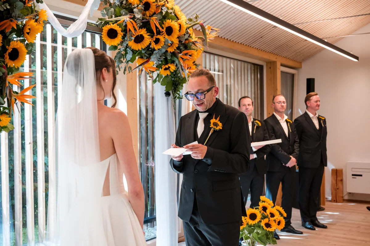The wedding couple during an indoor ceremony beneath a sunflower arch.
