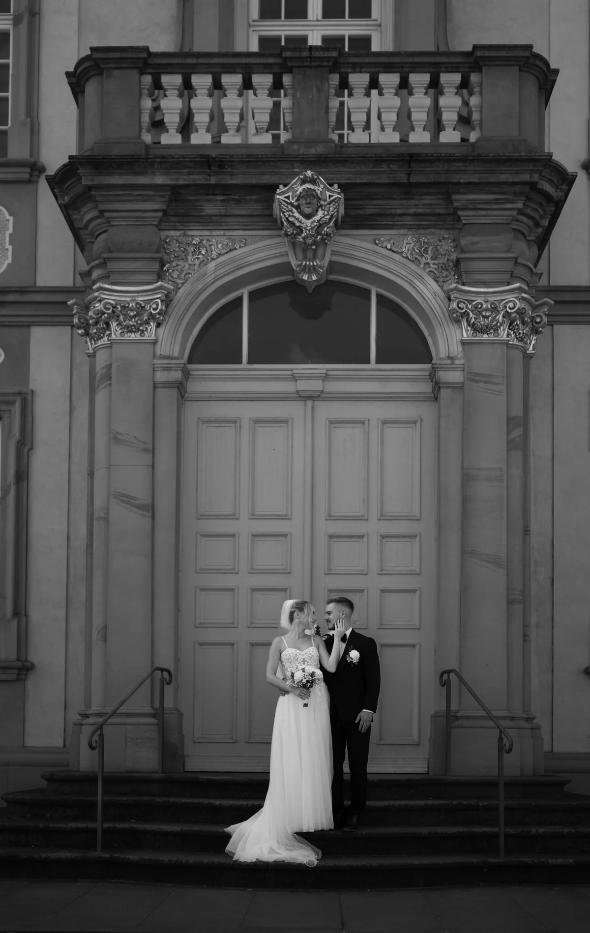 Black and white: the couple stand on the steps in front of a portal of Bruchsal Palace, with a balcony above.