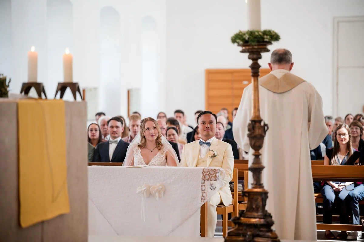 The couple sit at the front of the church looking at the priest, with the guests behind them.