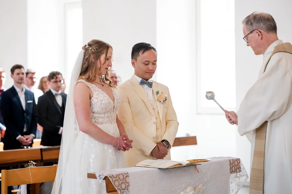 The priest blesses the couple with holy water as they stand at the altar with folded hands.