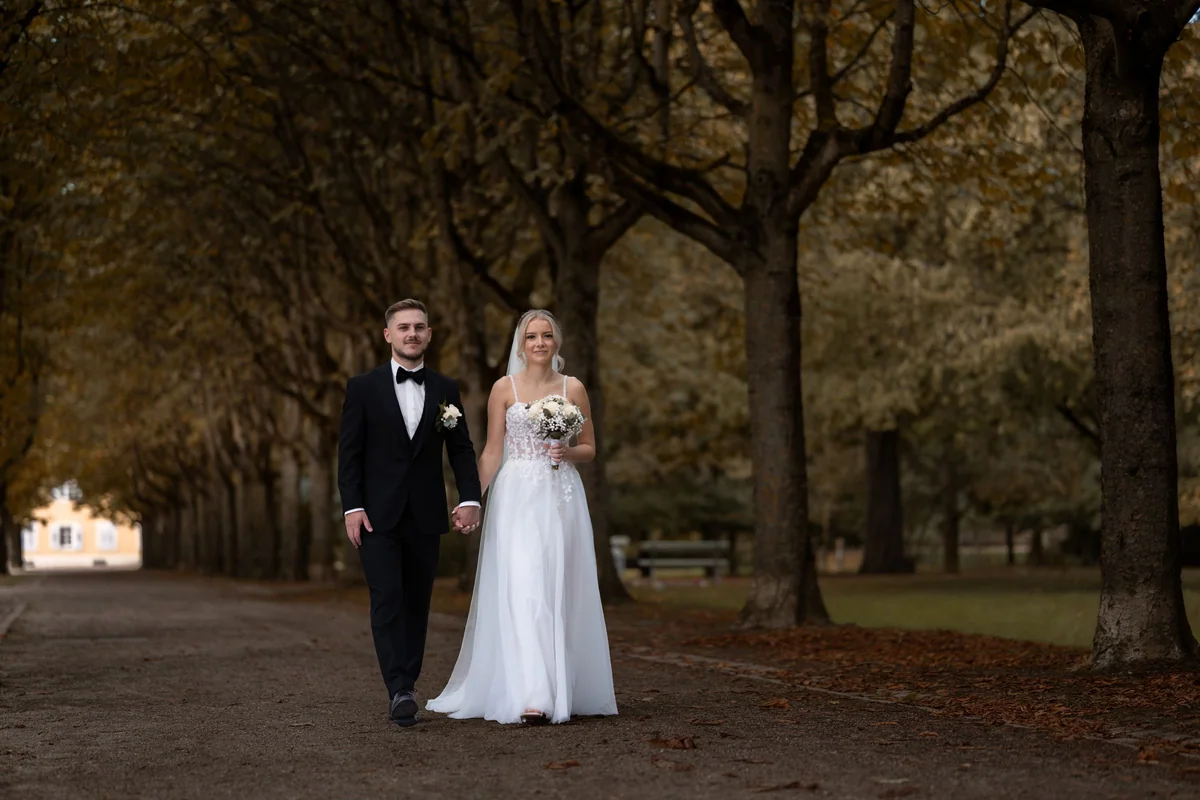 The couple walk hand in hand along a tree-lined avenue in the palace garden, autumn leaves on the path.