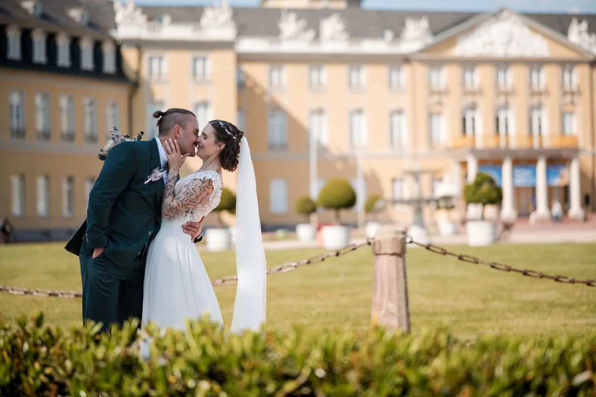 Ein Kuss vor dem Karlsruher Schloss, im Vordergrund Hecken und eine Kette.