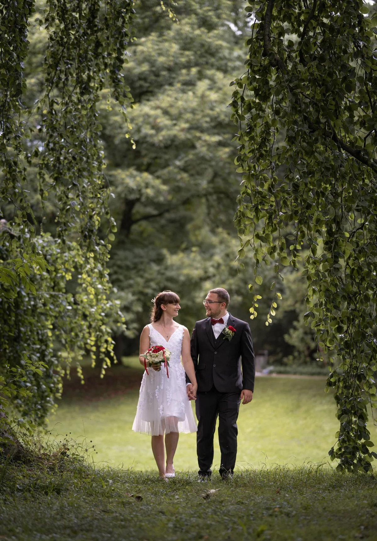 Ein Brautpaar geht Hand in Hand über die Wiese im Schlossgarten, eingerahmt von hängenden Zweigen.