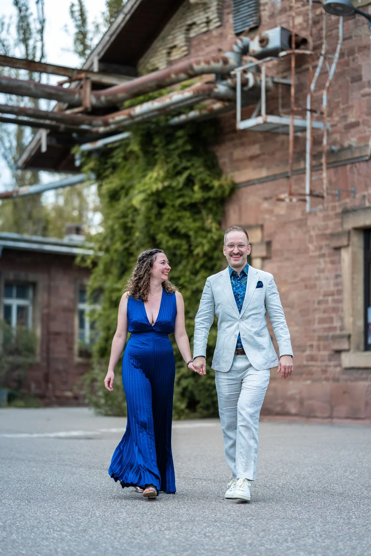 Ein Paar geht Hand in Hand an einem alten Backsteingebäude mit Rohren vorbei, sie trägt ein blaues Kleid.