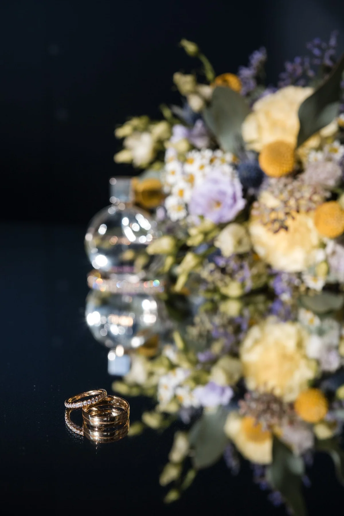 Blumen, Parfüm und Ringe spiegeln sich auf einer dunklen Tischfläche.