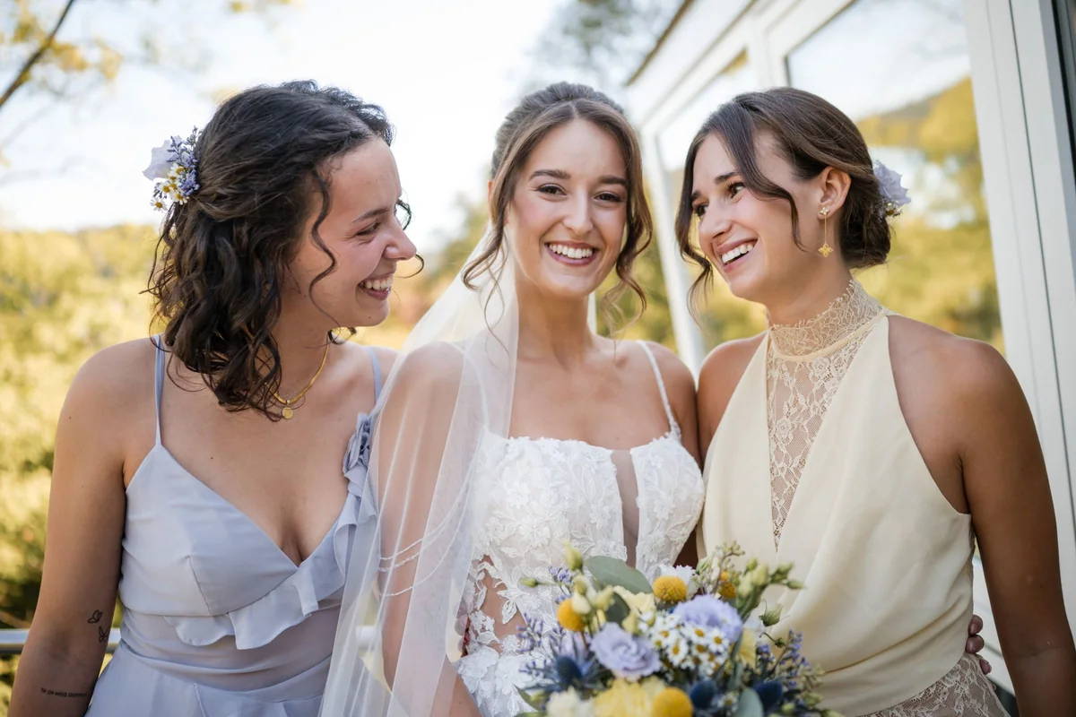 Drei Frauen lachen draußen zusammen, die Braut hält ihren Blumenstrauß.