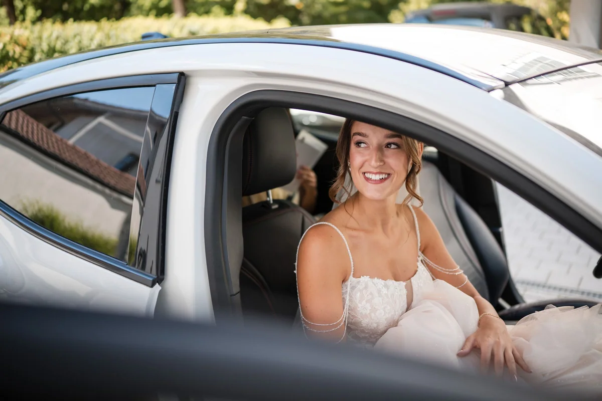 Die Braut sitzt lächelnd im Auto, ihr Schleier fällt über die Schulter.