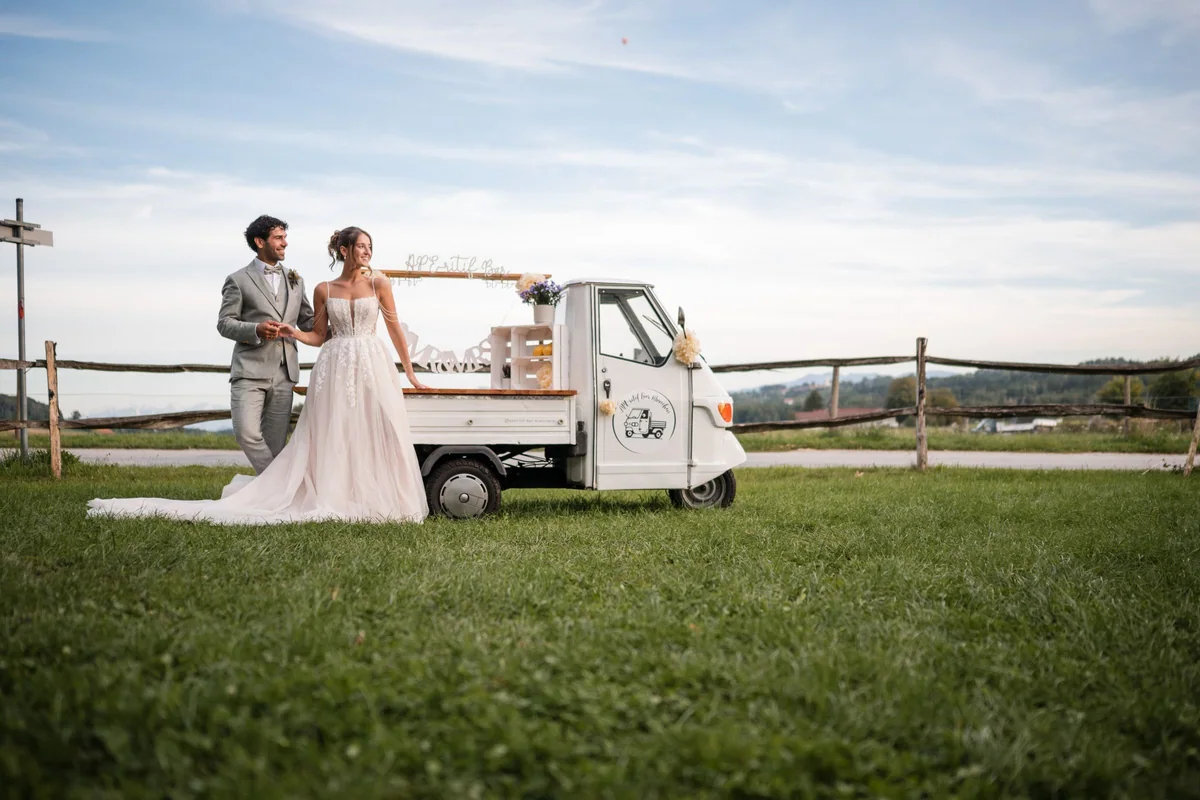 Das Hochzeitspaar lehnt an seiner weißen Ape vor der Landschaft von Baiern.