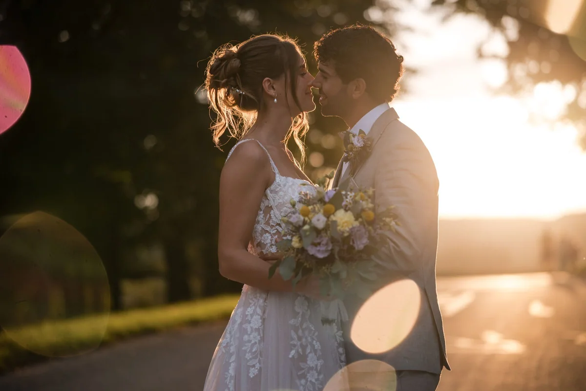 The couple kiss in warm backlight with a bouquet between them.