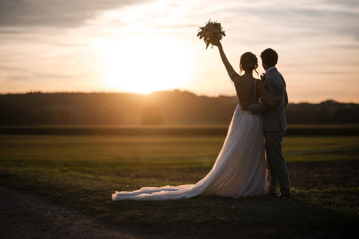 The couple stand in silhouette against the evening sky, holding up a bouquet.