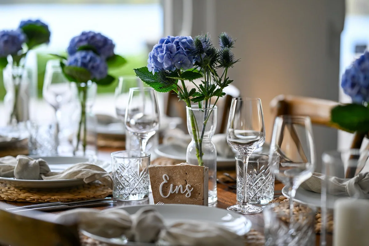 Laid tables with blue hydrangeas and crystal glasses in front of the large windows of the Seehaus.