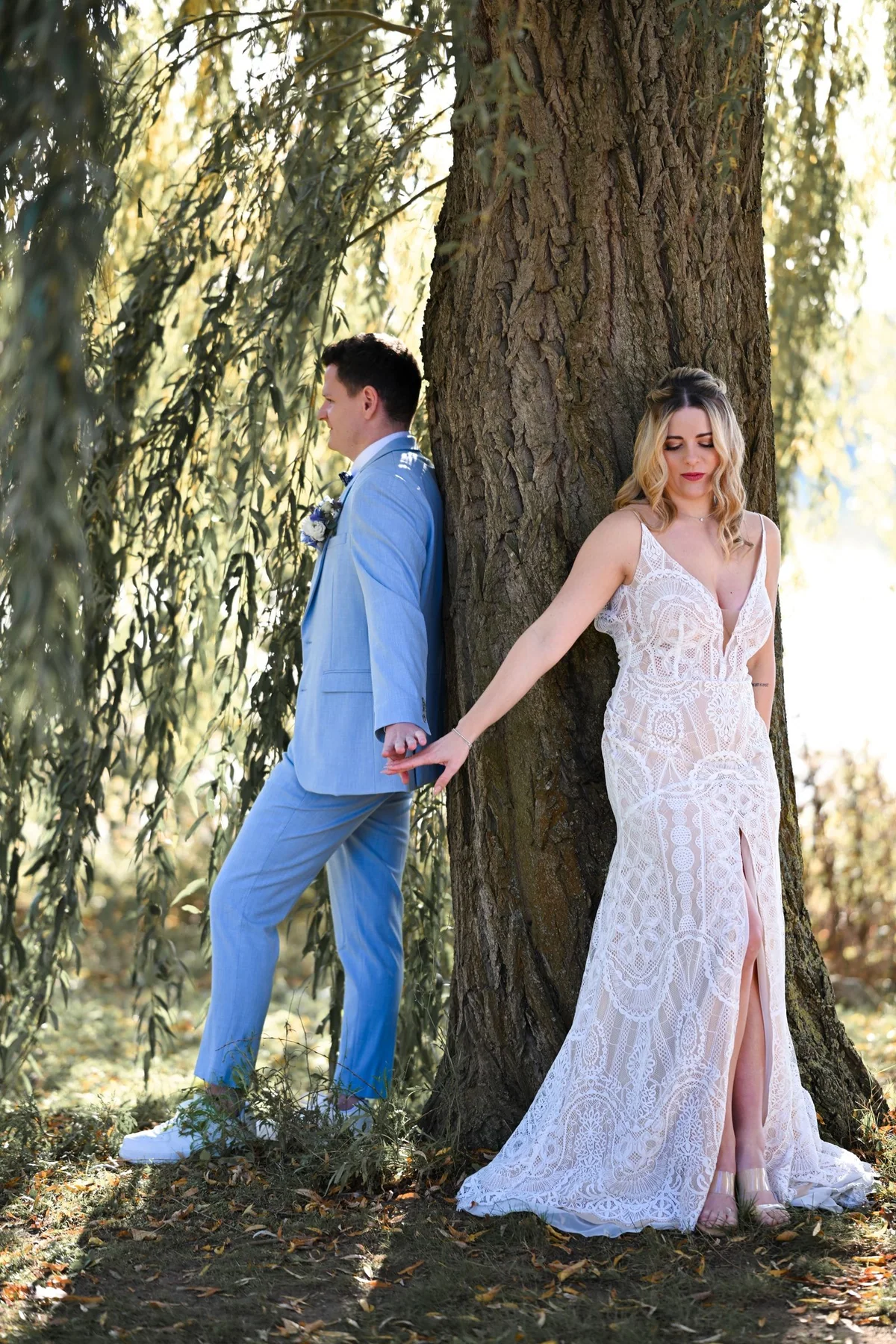 The bride and groom lean on either side of a thick tree trunk, holding hands.