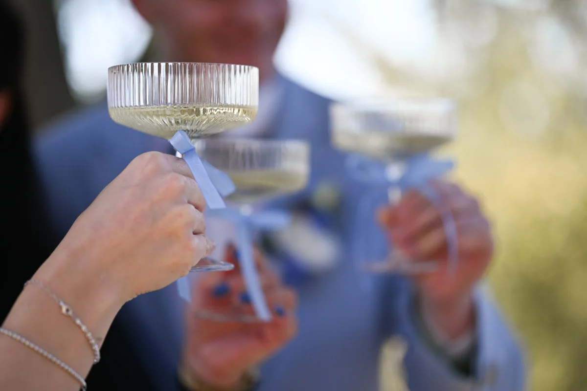 Two hands clink coupe glasses, light blue ribbons tied to the stems.