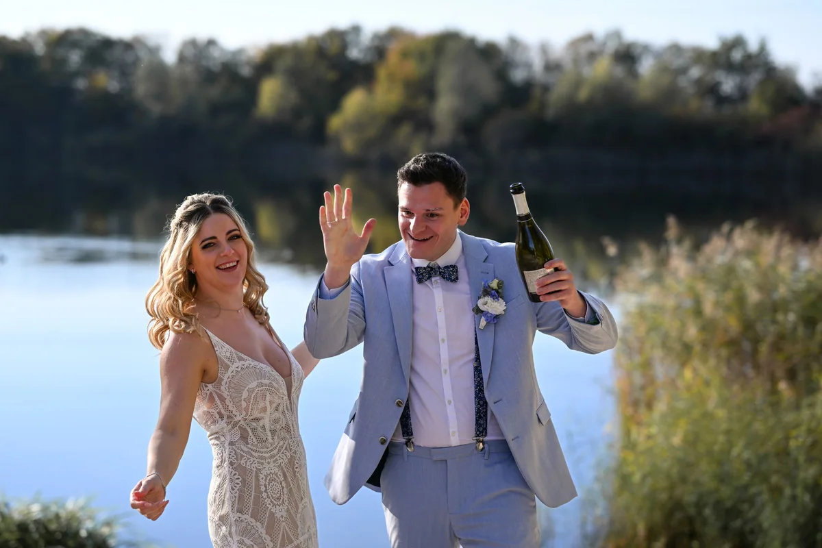 The bride and groom cheer on the lakeshore, he is holding a bottle of sparkling wine.