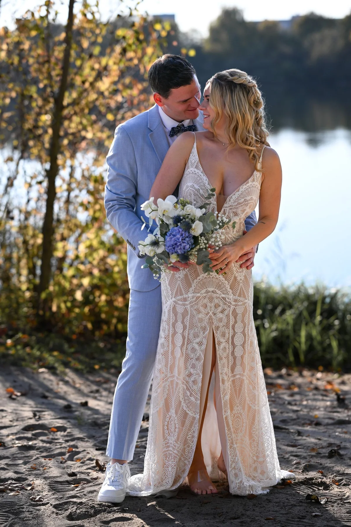 The couple stand in a close embrace on the sand by the shore, the bride holding her bouquet.