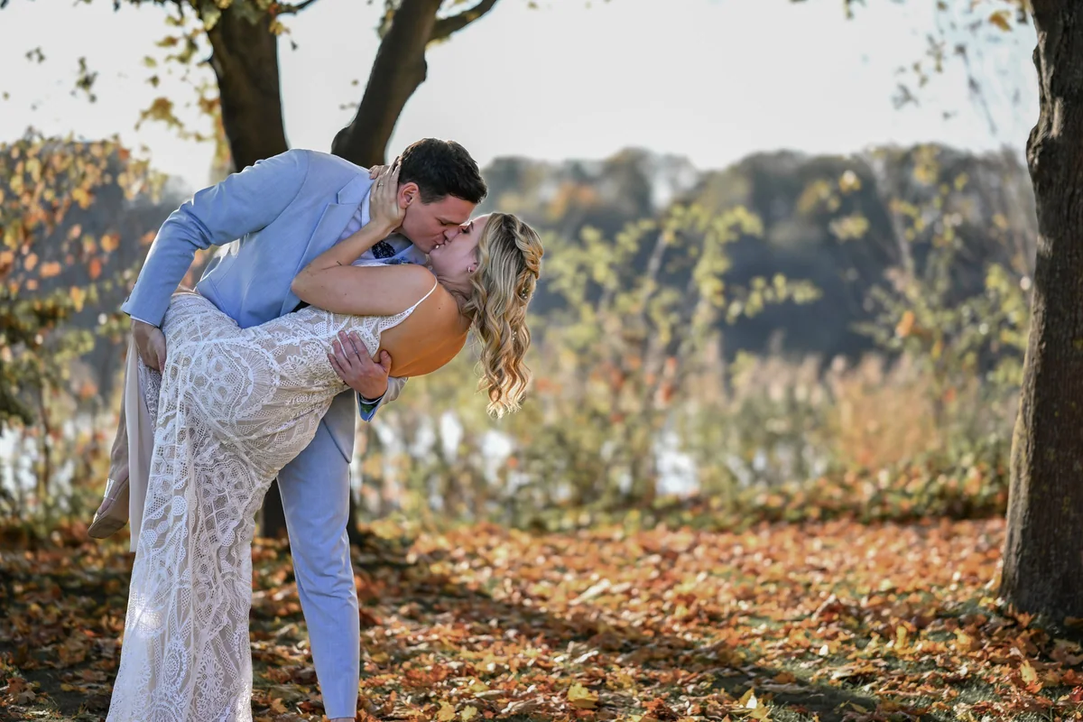 The groom dips the bride for a kiss, autumn leaves on the ground beneath the trees by the shore.