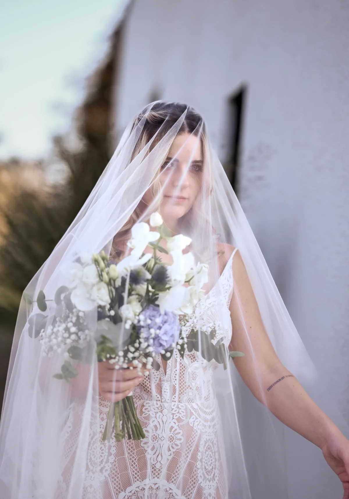 The bride stands beneath a long veil in front of the white wall, bouquet in hand.