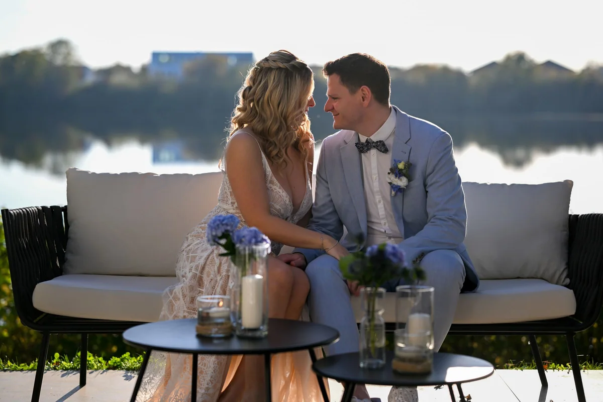 The couple sit on a lounge sofa right by the lake, with hydrangeas and lanterns on the table.