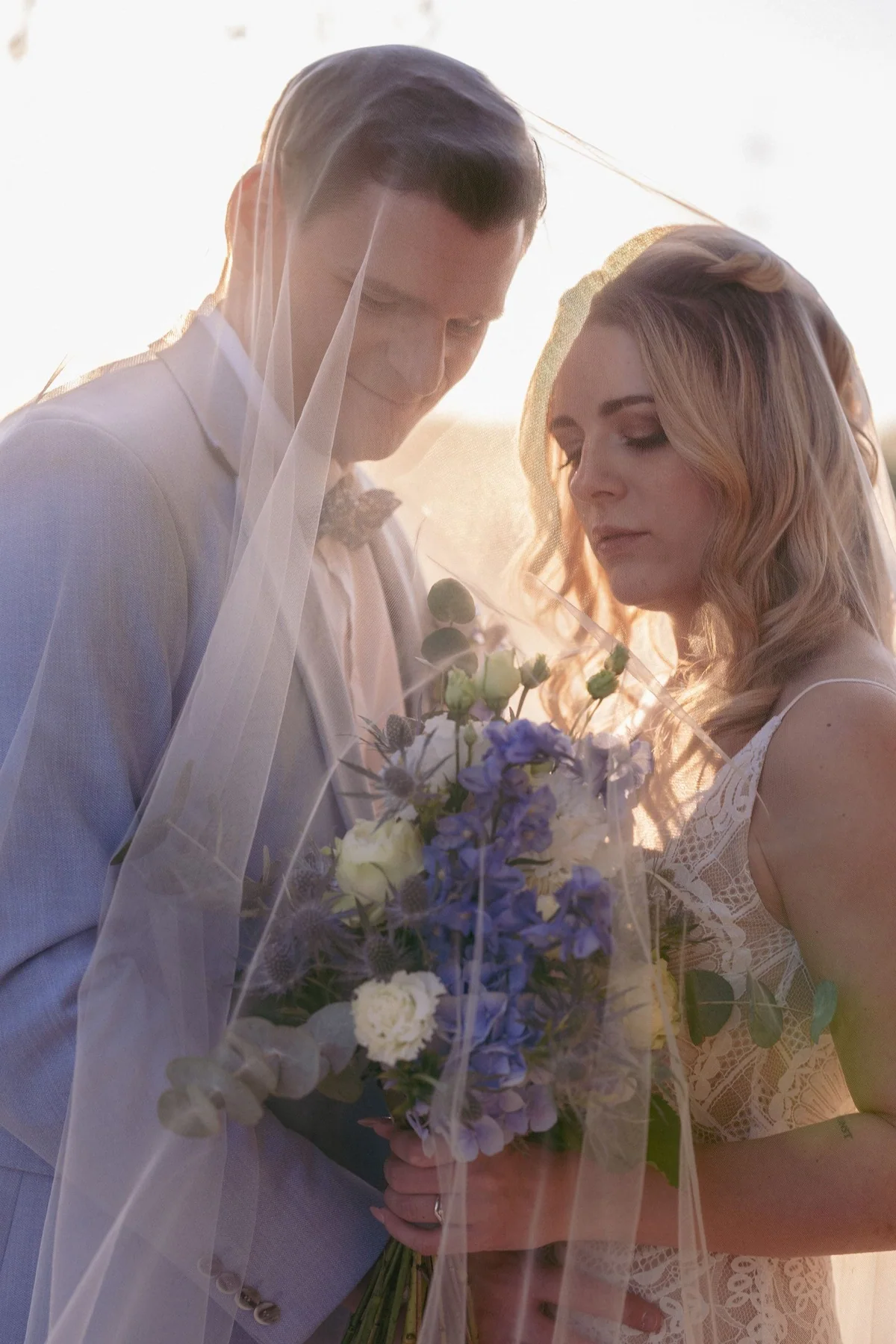 The couple stand beneath the veil in backlight, the bride holding a bouquet with blue flowers.