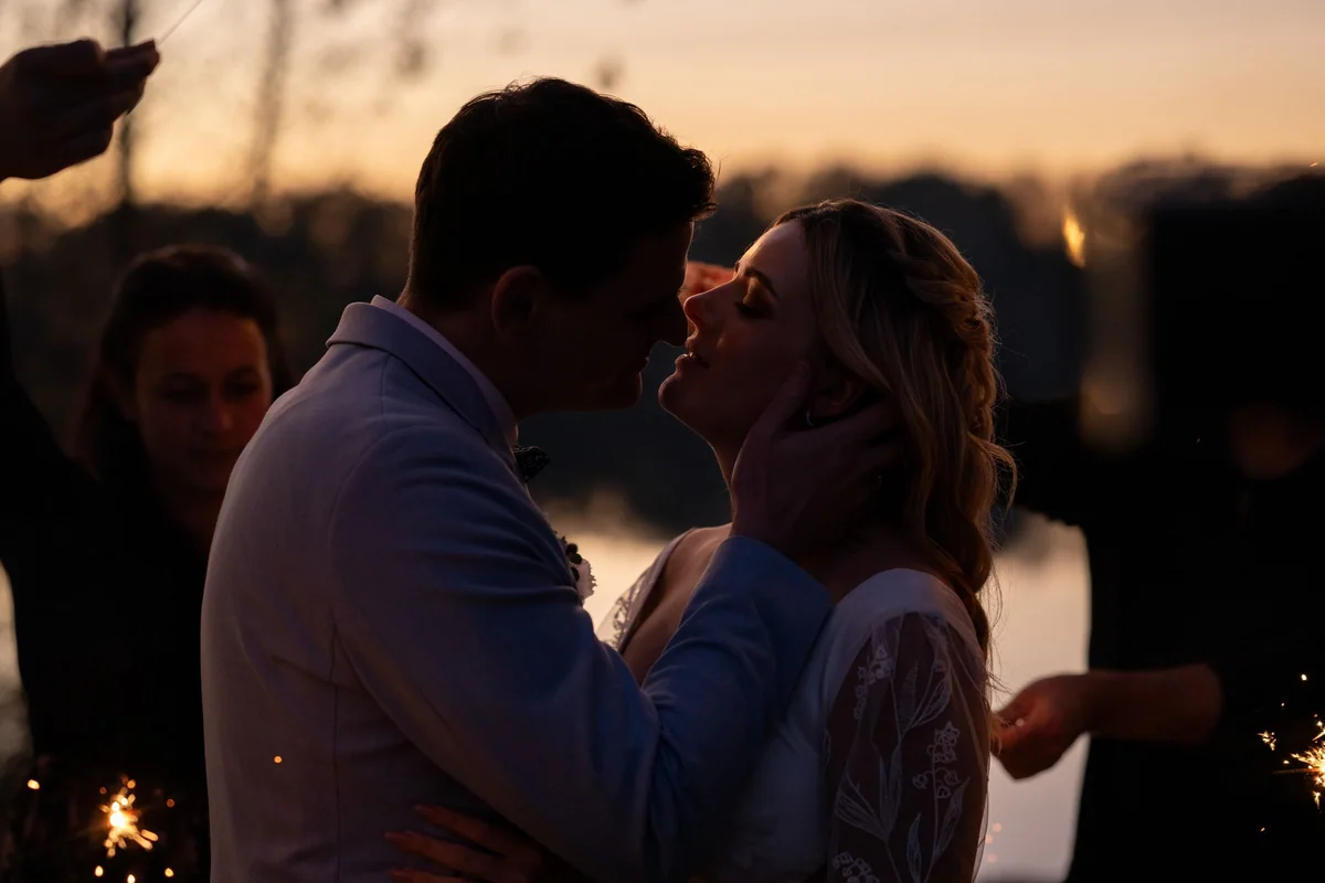 At sunset the couple kiss by the lake, surrounded by guests holding sparklers.