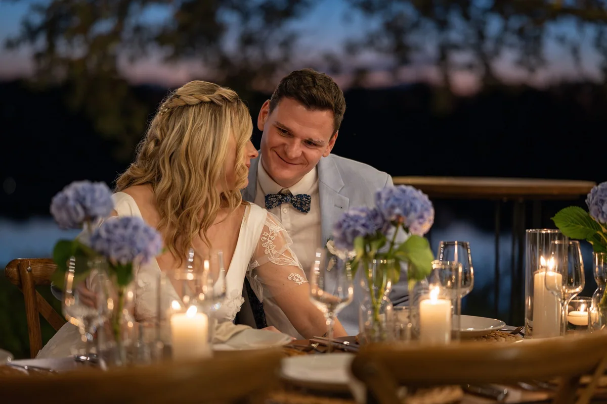 The couple sit laughing at the table in candlelight, the dark lake behind them.