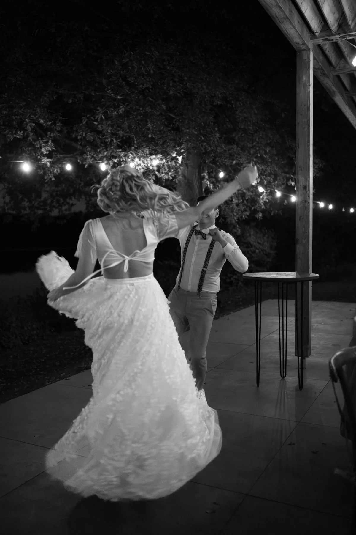 Black and white: the bride swirls her skirt while dancing, strings of lights glowing above.